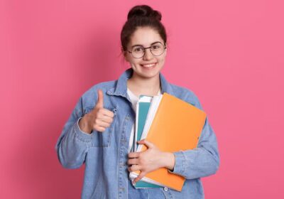 young-student-woman-wearing-denim-jacket-eyeglasses-holding-colorful-folders-showing-thumb-up-pink_176532-13861